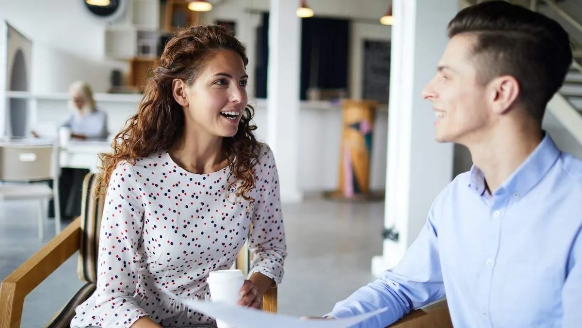 Two colleagues laughing together over a coffee and a document at a table